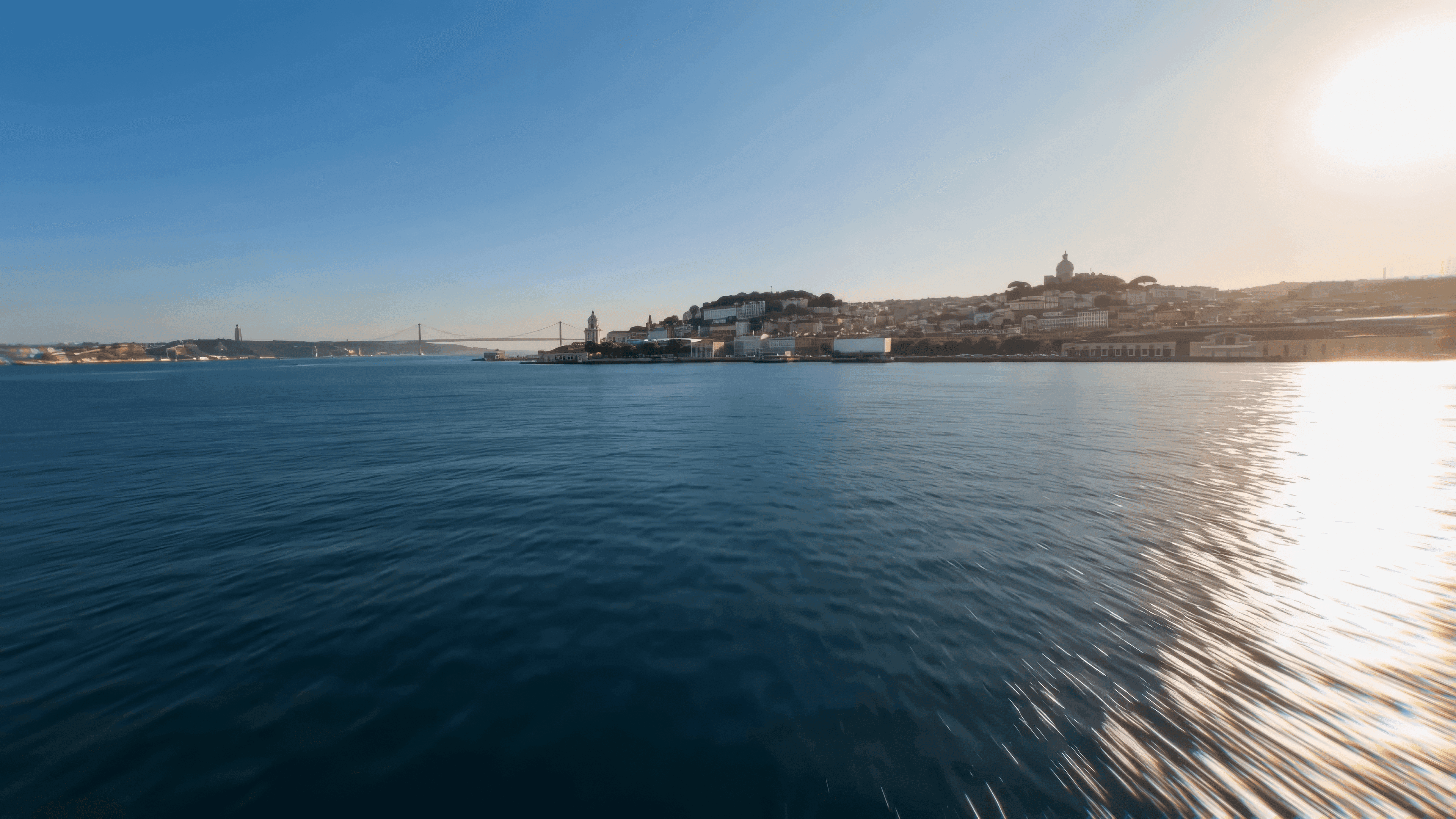 A city skyline rises from a sunny waterfront with a bridge and clear blue sky.